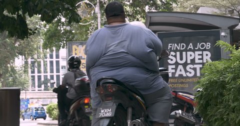 KUALA LUMPUR, MALAYSIA - CIRCA FEBRUARY 2018 - Extremely overweight man sitting on a motorcycle waiting for muslim women walking by before riding into city traffic