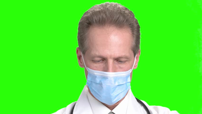 Mature doctor in medical mask smiling. Serious doctor in mask cheering up, green hroma background.