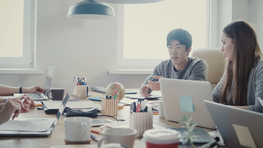 Happy Asian businessman takes a little globe, points at country at multiethnic office meeting in light loft coworking 4K