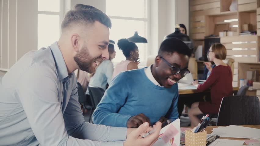 Two mixed race happy young men cooperate on business project in trendy stylish office, laugh, then get focused again 4K. | Shutterstock HD Video #1009815914