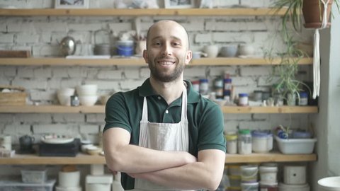 Portrait of male ceramist, who is standing in his studio and watching to the camera, slow motion. Young man in green polo t-shirt and white dickey has beard and smiling. Wooden shelves with colours