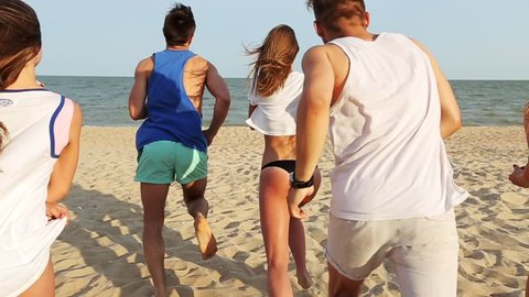 Back view of group of friends undress running into the sea water on sunset and throw shirts on the beach. Cheerful happy people having fun on the beach. Men and women go to swim in slow motion.