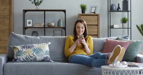 Beautiful young woman in the yellow sweater holding a smartphone in hands and typing on its screen while chatting in the living room at home. Indoors