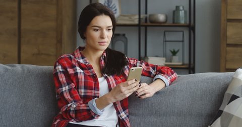 Pretty brunette young woman in the red motley shirt reading something on the smartphone and getting to know shoking news at home. Indoors