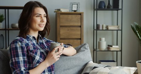 Attractive brunette young woman in the motley shirt drinking hot tea or coffee from a green cup on the gray sofa in the living room. Indoor