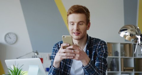 Caucasian young male startupper with red hair taping and typing on the phone while chatting and sitting at the table with a laptop in the modern office. Indoors