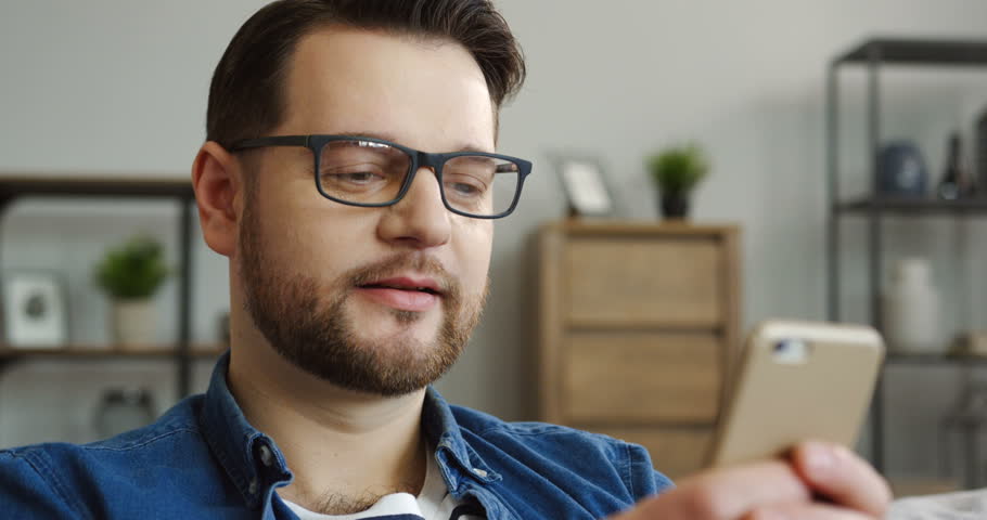 Portrait of the Caucasian middle-aged man in the jeans shirt typing and chatting on the smartphone while sitting in the living room. Indoor
