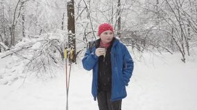 Caucasian teenager in winter forest drinks hot tea at short break during cross-country ski walk. There is heavy snowfall. Boy looking around