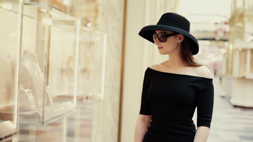 A beautiful stylish lady in a hat and sunglasses approaches the shop window of a boutique and looks at the goods with interest. Luxury jewelry store | Shutterstock HD Video #1009843763