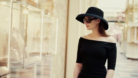 A beautiful stylish lady in a hat and sunglasses approaches the shop window of a boutique and looks at the goods with interest. Luxury jewelry store