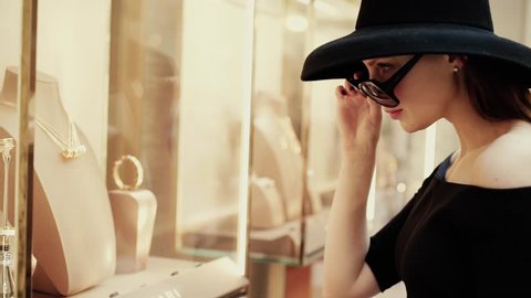 A beautiful young stylish rich girl in a hat takes off her glasses to see the goods on the shop window of a luxury boutique. Slow motion