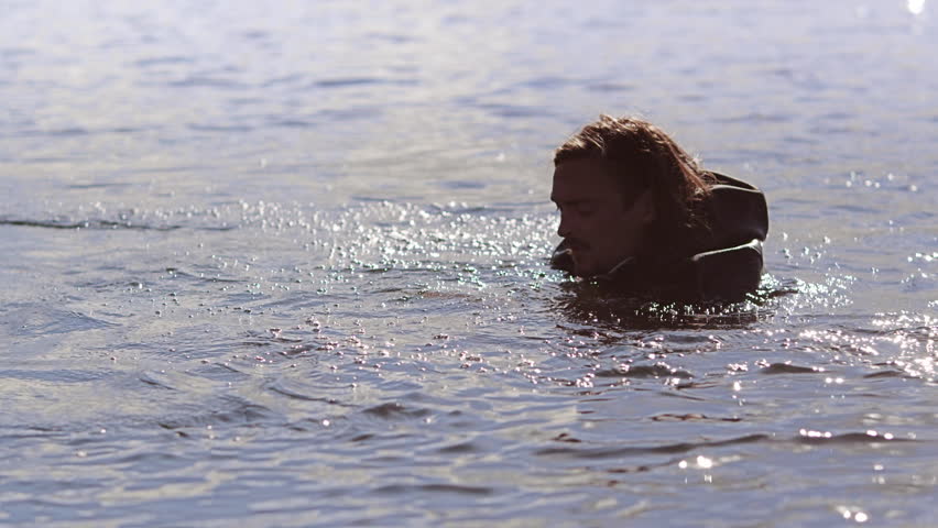 Man in waterproof clothing preparing to ride on wake board. Young man splashing in water in swimming suit. Sportsman in water before start