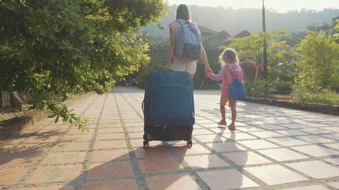 Steadicam shot of young mother with little daughter arriving at hotel, goes with suitcase on the beautiful garden toward lobby for checking in, slow motion.