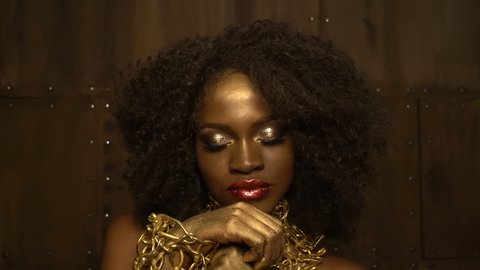 Charming african woman with big red glossy lips, golden eyeshadows and dark curly hair wearing the golden chain bracelets is looking in camera at the brown background. Video stock