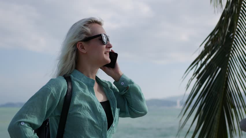 Happy caucasian woman with long blonde hair in sunglasses and green shirt standing and talking on phone near palm tree on a blue sky and sea background. Travel concept