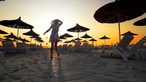 Woman in white dress and hat walk toward camera on beach in the seaside resort with sun loungers and wooden sheds against sunset, cinematic