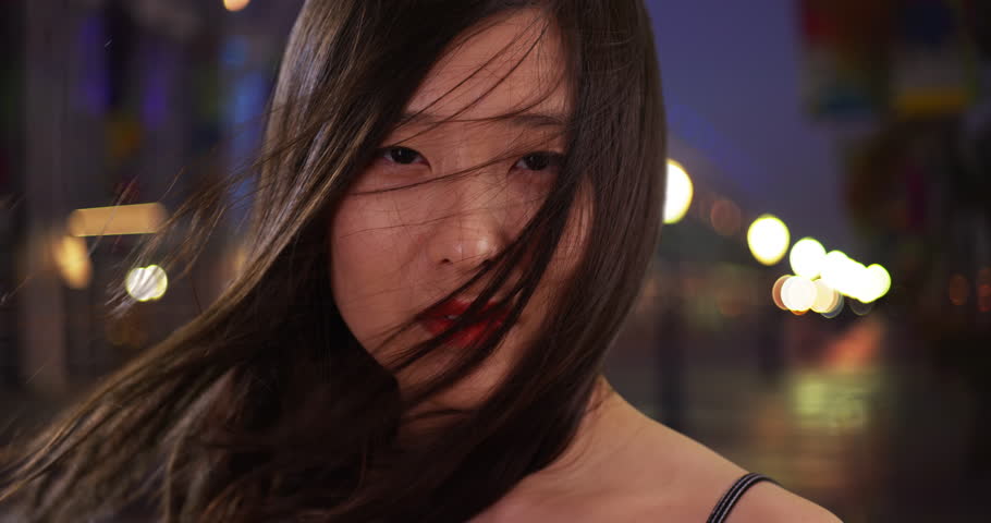 Close up of lovely millennial woman with hair blowing in the wind in urban setting. Portrait of Asian female looking at the camera in night scene. 4k