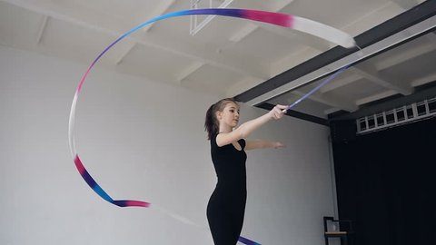 Attractive female gymnast dancing with colored ribbon in gymnastics school. Slim gymnast in black body performs the gymnastics exercise with a ribbon in the white studio and possing to the camera