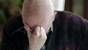 Elderly man overcome with grief sitting with glasses in hand and head bowed before looking up crying.