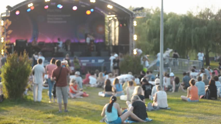 Celebratory concert and watching people outdoors on summer day. Singing artists on Irrl stage, men women stand and sit on grass in warm weather, next to green trees. Festive event in open air people Royalty-Free Stock Footage #1009909508