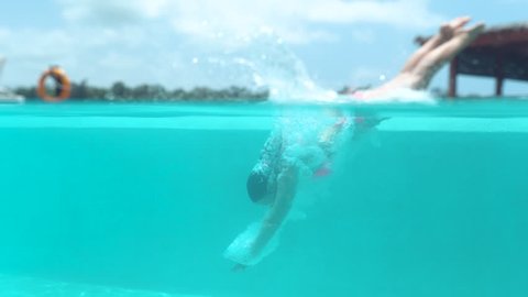 SLOW MOTION, HALF UNDERWATER: Active young woman dives into empty pool and splashes glassy water. Caucasian girl jumps head-first into refreshing pool during relaxing stay in tropical hotel resort.