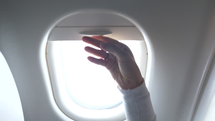 CLOSE UP: Young woman looks through the airplane window and closes the blinds. Female traveler on transatlantic flight watches the bright sky and the large airplane wing before closing the shades.