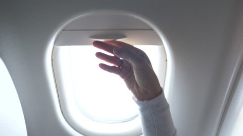 CLOSE UP: Young woman looks through the airplane window and closes the blinds. Female traveler on transatlantic flight watches the bright sky and the large airplane wing before closing the shades.
