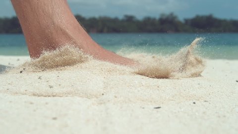 SLOW MOTION, CLOSE UP, LOW ANGLE: Unknown man steps barefoot into scorching hot white sand on idyllic exotic beach. Male foot steps into sand and makes grains of sand fly around calm tropical coast.