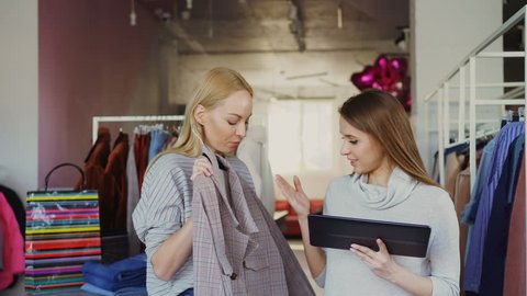 Young business owner is using tablet while standing in her clothing store. Her assistant is coming with garment and asking advice. Businesswoman is showing her where to place it.