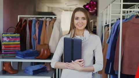Portrait of young businesswoman standing in her clothing boutique, holding tablet, smiling and looking at camera. Spacious store with women's clothes in background.