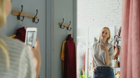 Pretty blond woman is making mirror selfie with smart phone while standing in fitting room in clothes boutique. She is posing, moving and smiling carelessly.