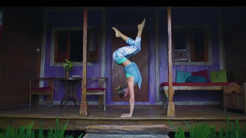 Woman in yoga sports clothing doing handstand yoga asana in front of a wooden house in bali