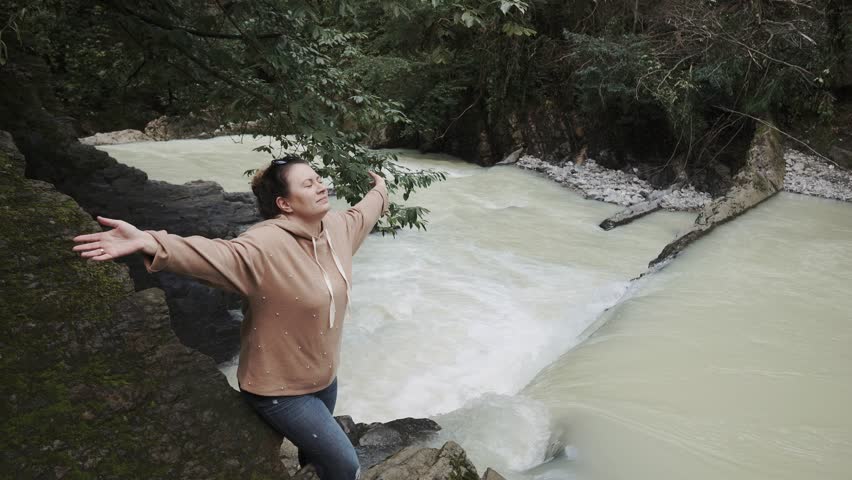 a young woman in warm clothes spread her hands to the sides and enjoys the drops of water from the waterfall that shine her, the lady enjoys the natural beauties