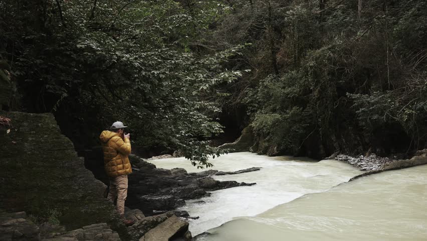 Happy and cheerful tourist with a camera taking pictures of river in mountain. He is wearing warm clothes outdoor.