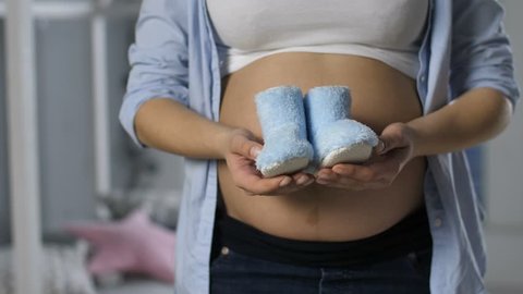 Close-up of pregnant female with big naked belly showing blue baby shoes for newborn boy to camera. Happy expecting mother offering sweet infant booties
