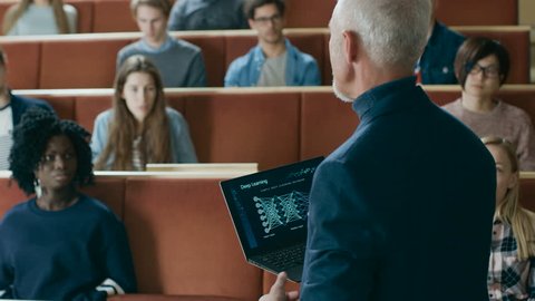 Back View of the Professor Giving Lecture on Computer Science to a Classroom Full of Multi Ethnic Students. Teacher Holds Laptop with Deep Learning, Artificial Intelligence Infographics on the Screen.