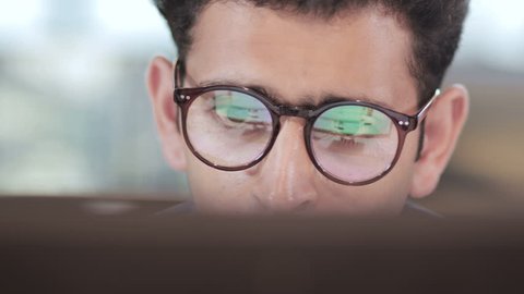 close up of a young and busy businessman wearing eyeglasses working on computer. Reflection of computer screen can be seen in the eyeglasses of an handsome male entrepreneur in modern corporate office