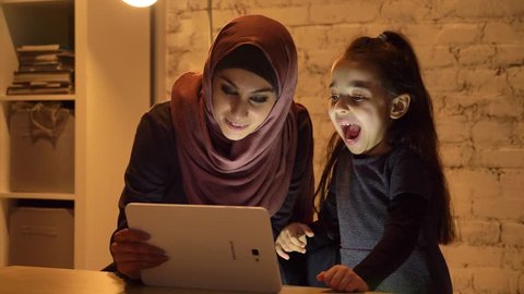 A young mother in a hijab sits with her little daughter on the couch in the evening and uses a tablet, watch a funny video, laughing, home comfort in the background 50 fps 库存视频