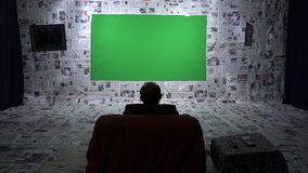 Over the shoulder shot of a man sitting and then stand up, go to the bigger LCD television with a green screen
