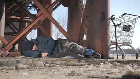 Full length cold homeless beggar male in jacket sleeping under the bridge in fall, eyes covered with hat. Urban homeless person with no home staying overnight outside. Dolly shot