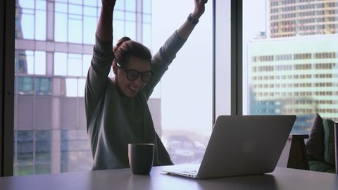 Business woman with arms raised is celebrating success. Attractive female freelancer in glasses is smiling and dancing in office