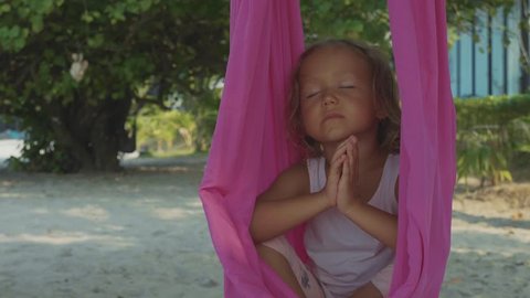 Little cute child girl doing yoga exersice with namaskar gesture hammock on the beach in slow motion.
