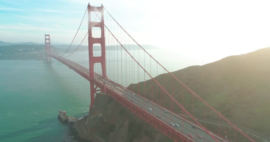 Aerial view of the Golden Gate landmark with San Francisco green hills on a background. Drone flight outbound the tower of the red bridge with the sunset view above the Pacific Ocean, West coast. 4K