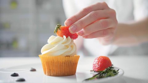 Dessert cook female hand decorates tasty cupcakes white cream topping with cutted strawberry pieces at kitchen