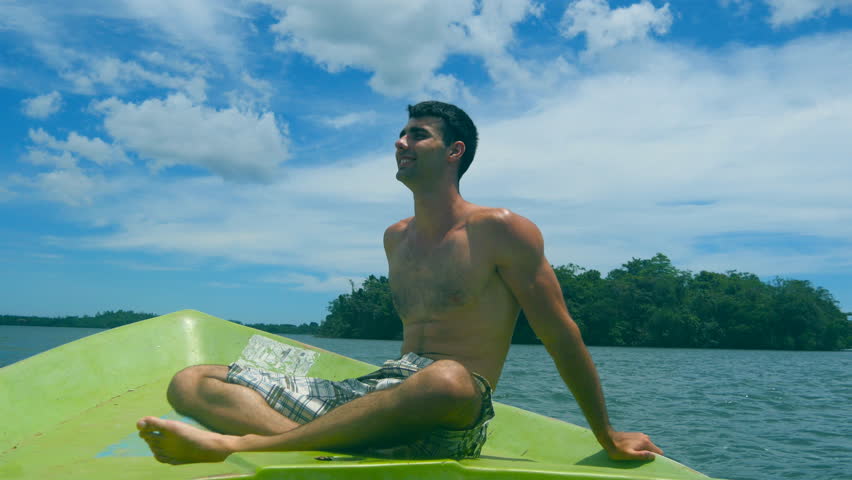 Side view of young athletic guy sitting on bow of boat and looking to beautiful nature landscape on sunny day. Happy sporty man smiling on deck of ship and enjoying summer trip. Close up