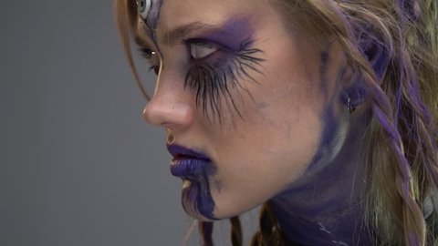 A girl with a third eye in her forehead and purple streaks near the eyes and lips looks into the camera, slow motion