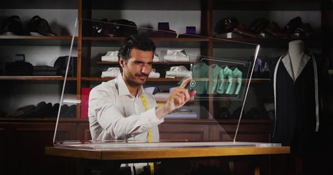 A man in a tailor shop, he clicks a glass monitor to select the material and the type of shirt to be bought with the holography. Concept of: future, technology and tradition.