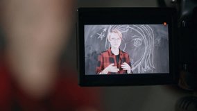 Shot through display of video camera of teenage girl in glasses standing before blackboard with drawing and gesturing while talking to her viewers