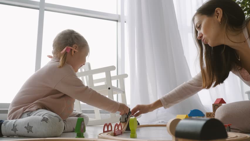 Close-up of little cute girl and her mother playing with wooden toy railway while sitting on the floor in white living room | Shutterstock HD Video #1010060846