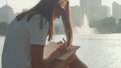 Young woman sitting on the bench in park and writing in diary, close-up. Woman write morning pages at park with lake in megapolis with skyscrapers on the background. Stockvideó